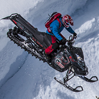 A man in a blue jacket and red pants dropping a black sled over a cornice.
