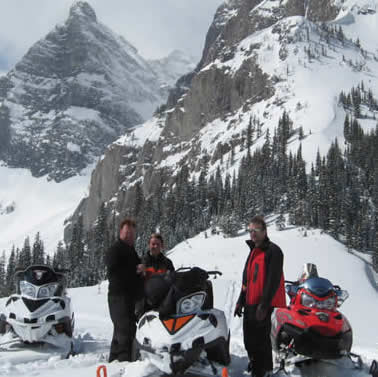 Two men, a woman and four snowmobiles stand on snowy ground before a stark mountain range 