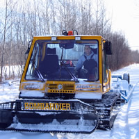 A yellow groomer comes down a trail with trees on both sides. Danny Smyl is driving.