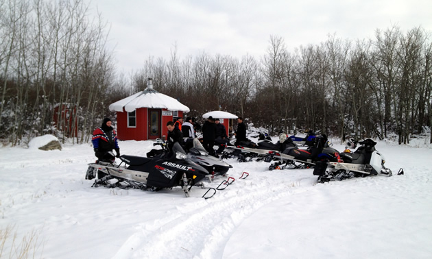 A group of sledders gather around a short, red, round building surrounded by bush.