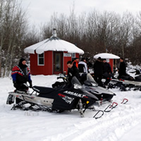 A group of sledders gather around a short, red, round building surrounded by bush.