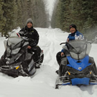 Three snowmobiles and two riders on a snowy trail among trees