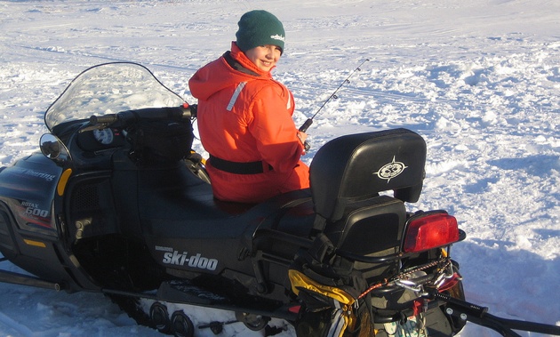 A youth enjoying ice fishing while sitting on his snowmobile.