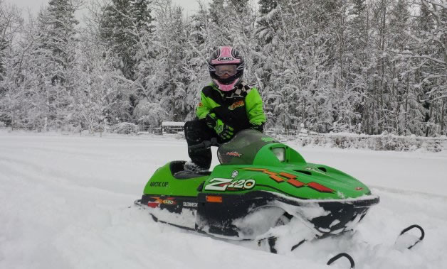 A small girl standing on a green Kitty Kat snowmobile. 