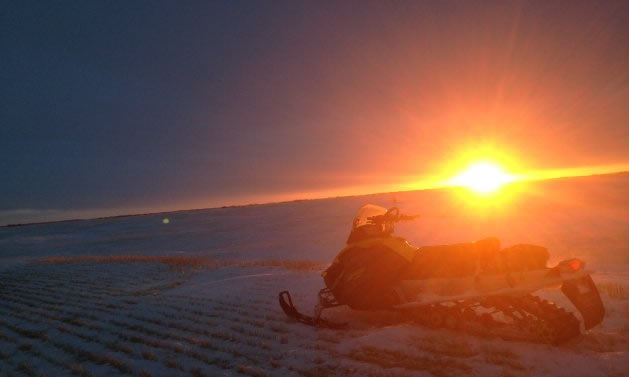 A snowmobile parked in the prairies with a beautiful sunset behind it. 