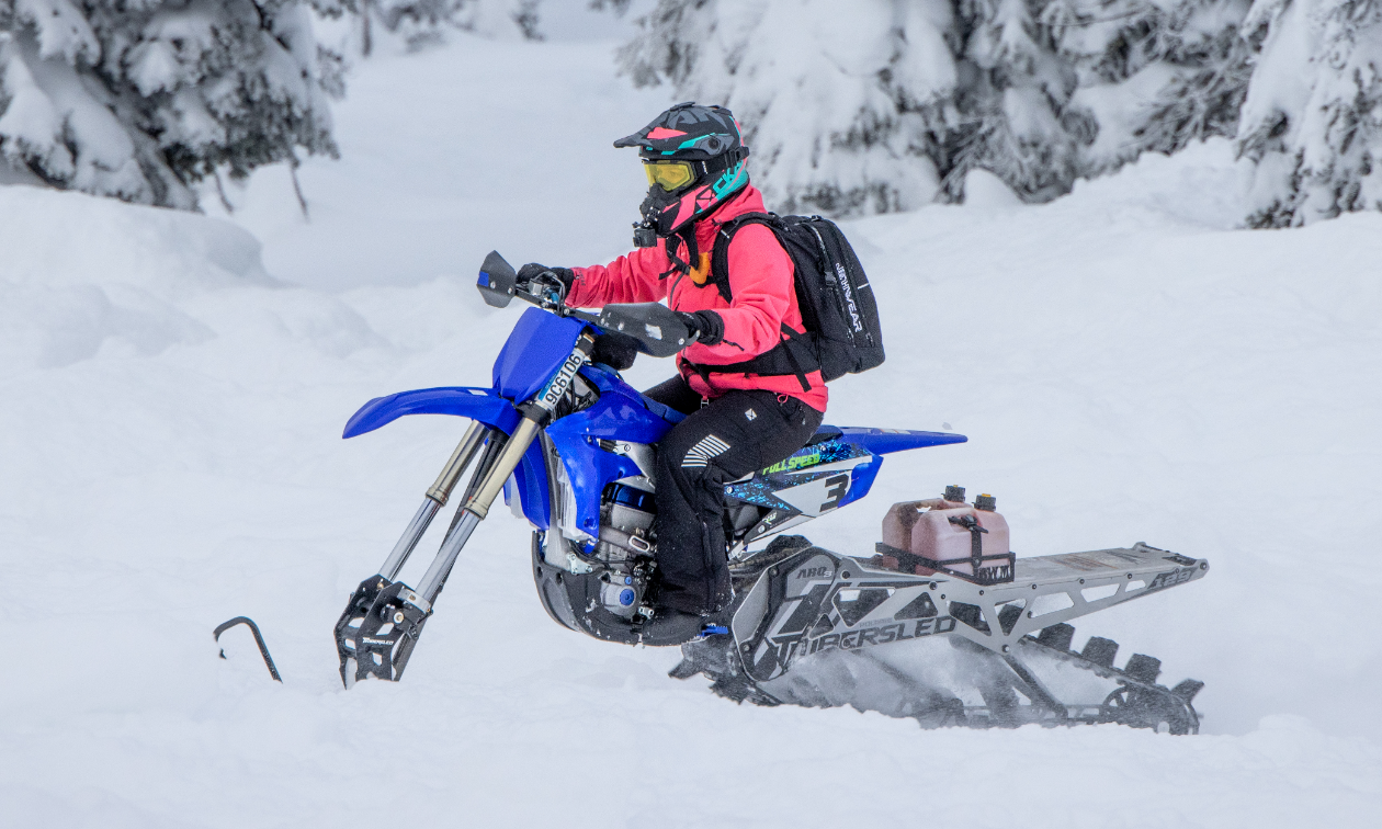 Cassandra Mainville wears a pink jacket and sits on a blue snow bike in a snowy forest. 