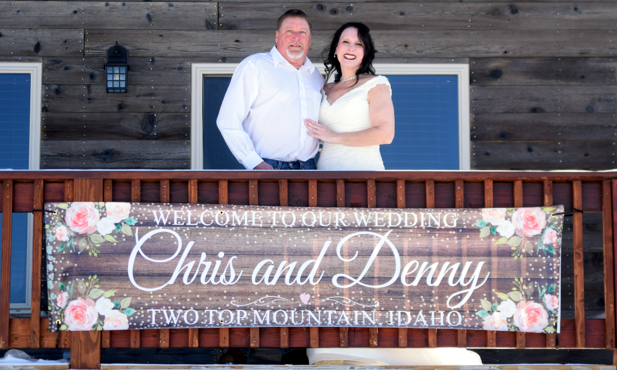 Denny and Chris Steigerwald stand on a cabin balcony in front of a sign that says Welcome to our wedding: Chris and Denny, Two Top Mountain, Idaho