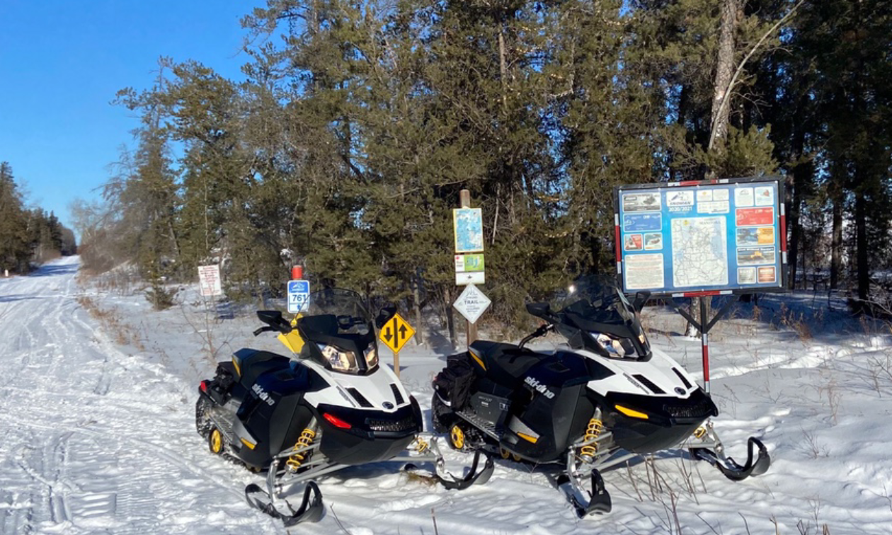 Two snowmobiles are parked in front of road signage next to a groomed trail.