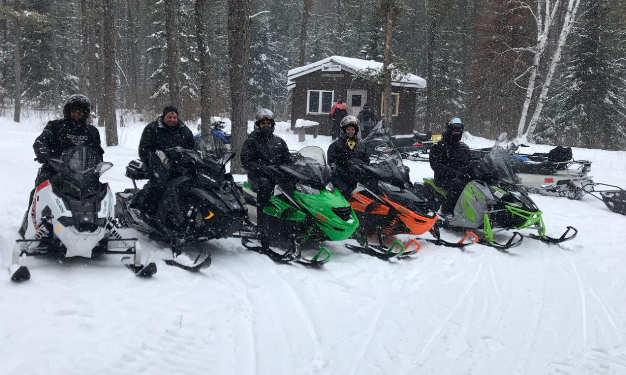 Five snowmobilers pose for a photo in front of a warm-up shelter as snow falls.