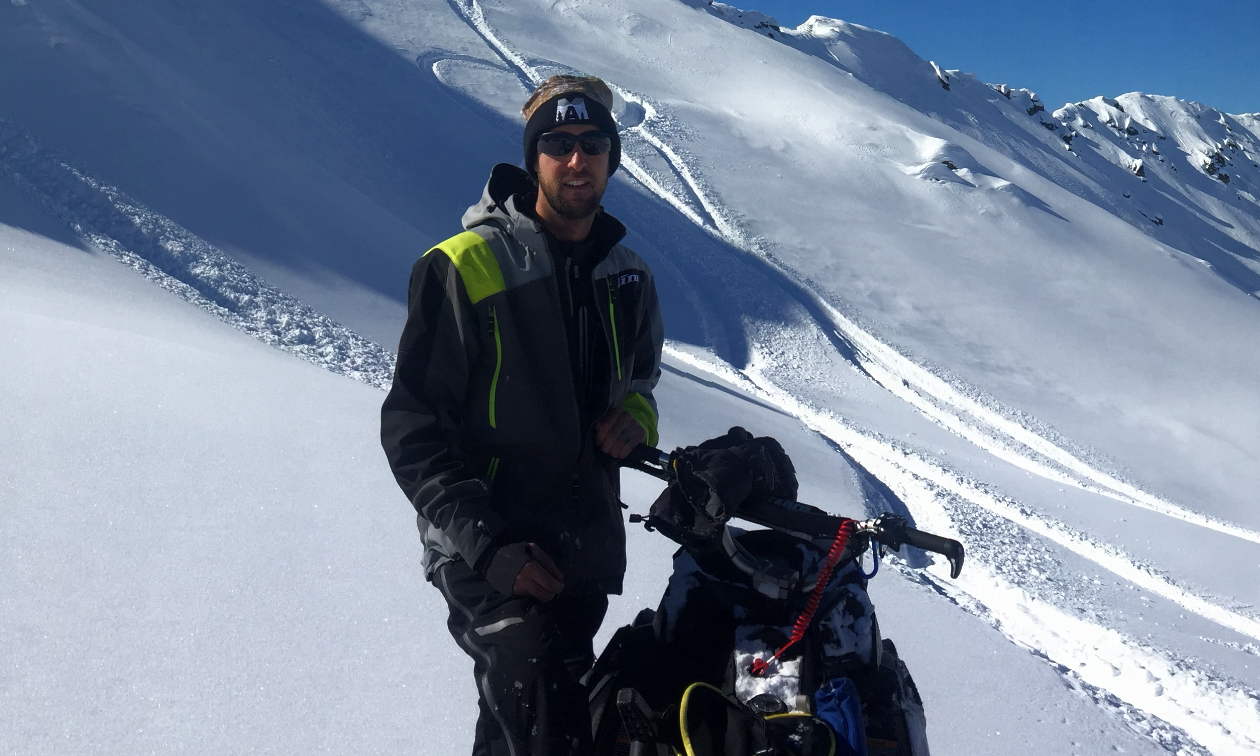 Sheldon Kelly stands next to his snowmobile on a calm mountain filled with snow. 