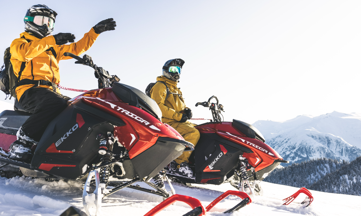 Two snowmobilers with yellow jackets sit on red Taiga snowmobiles on top of a mountain. 