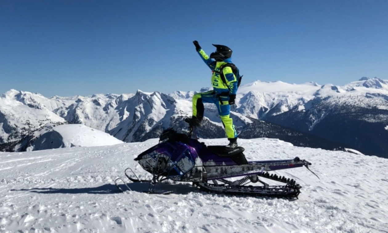 Andrew McKenzie stands on top of his black snowmobile, holding up his hand with a skyline of mountains in the distance. 