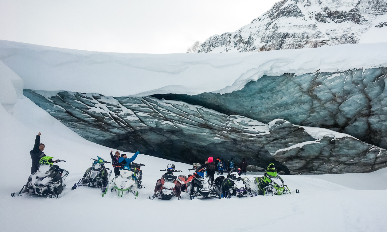 A row of snowmobilers pose in front of a glacier. 