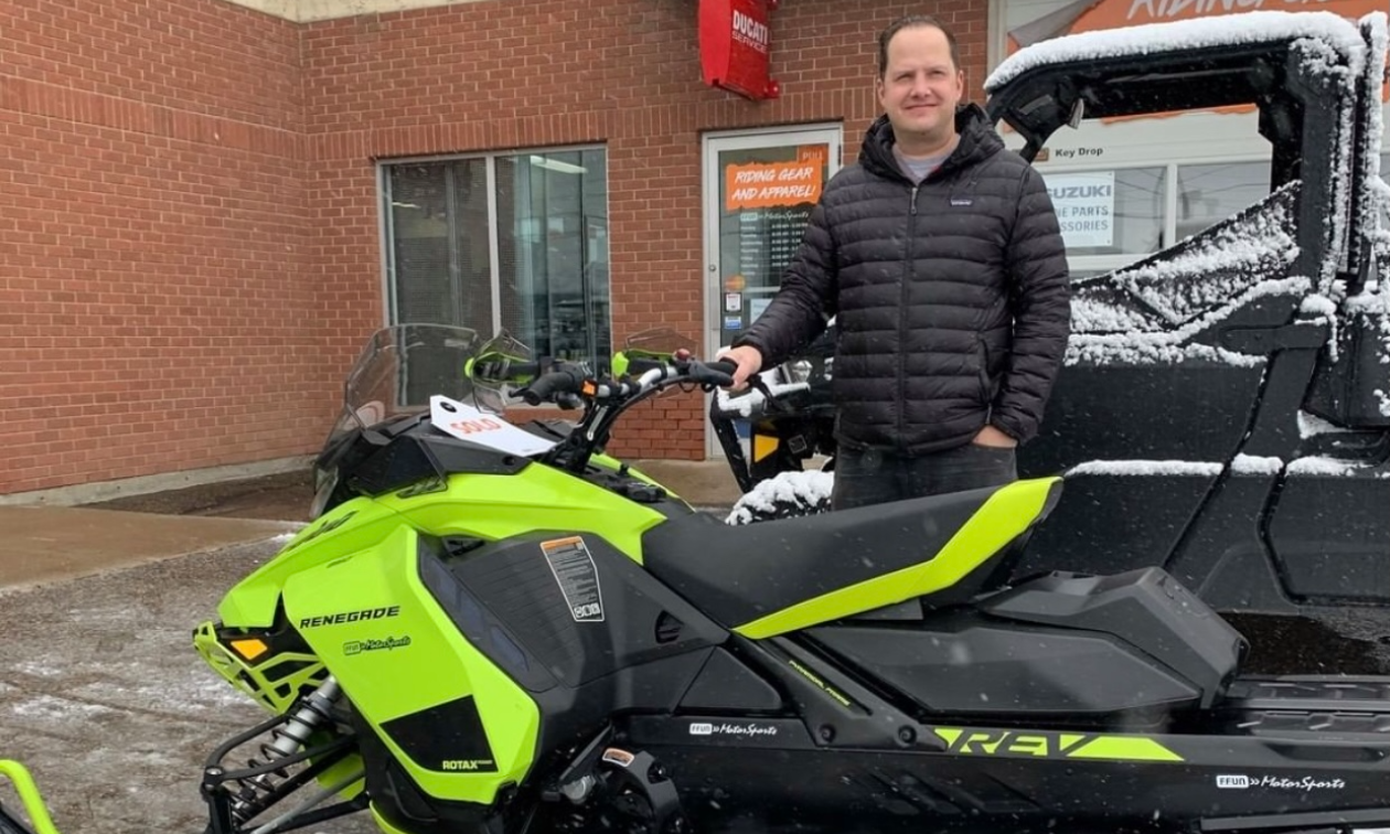 Audun Klath stands behind a green snowmobile in a parking lot. 