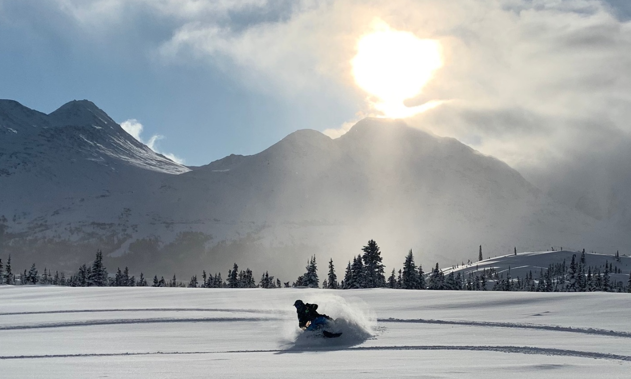 A snowmobiler cuts through snow in a glad as the sun shines above the mountains in the distance. 