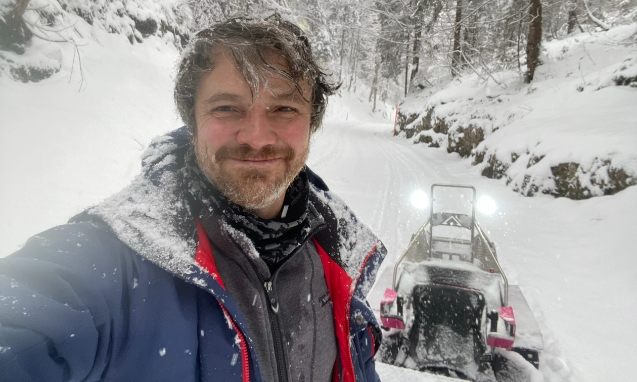 Sergey Ignatyev smiles amidst falling snow in a big blue jacket in front of his parked Bobsla electric snowmobile. 