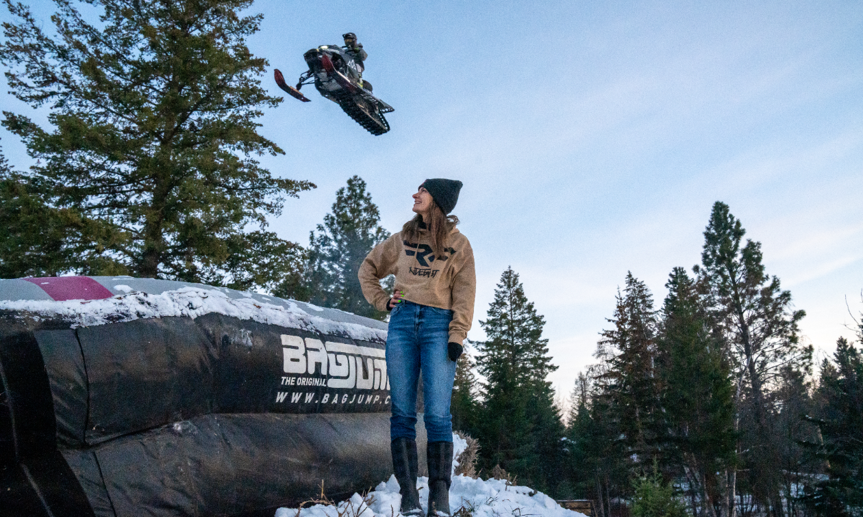 Brandon Milford soars into the air on his snowmobile while a woman looks on next to an inflatable landing platform. 