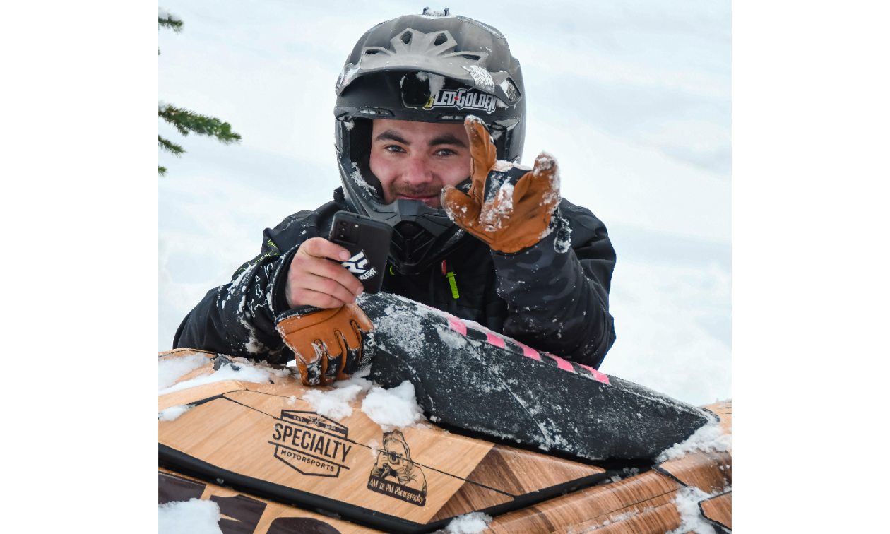 Brandon Milford sits in the snow behind his snowmobile and holds up the horns using his index and pinky fingers on his gloved left hand. 