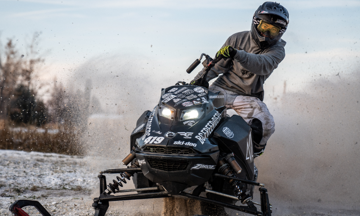 Brandon Milford looks into the camera from close distance while he twists his snowmobile, spraying dirty snow behind him. 