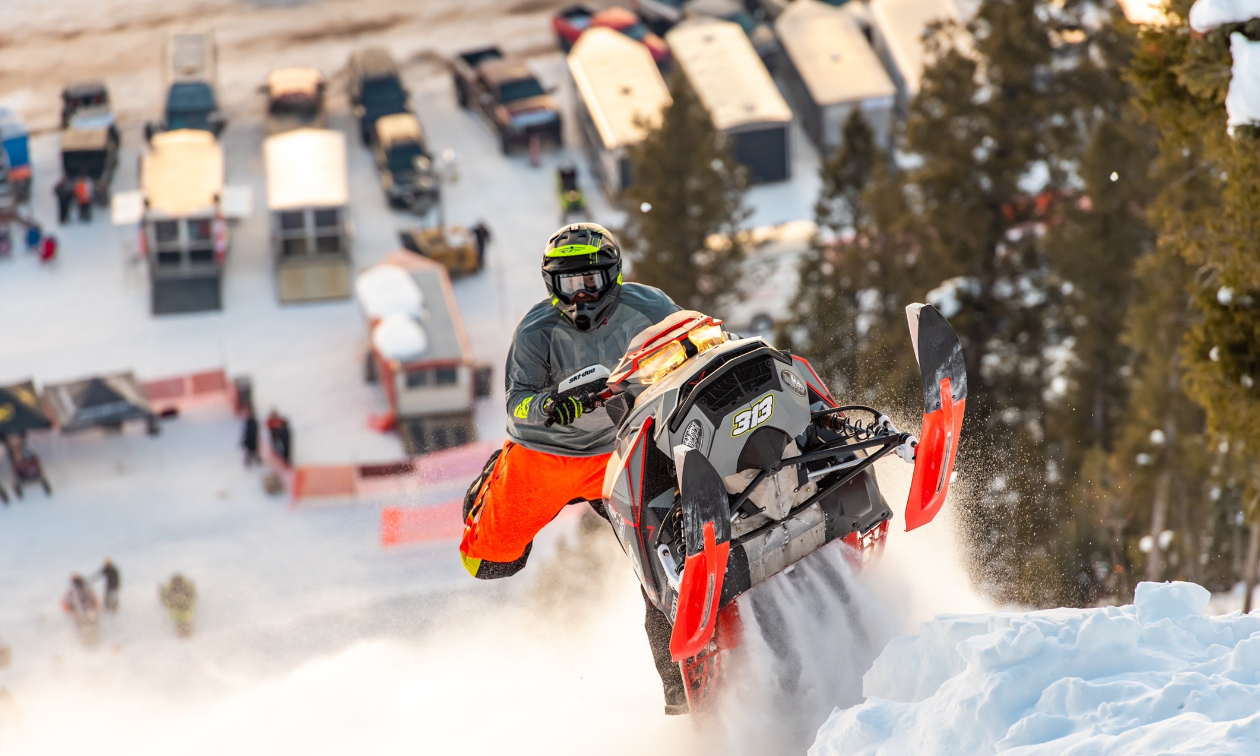 Brennan Mulvahill hill climbs his orange and grey snowmobile up a mountain with trailers at the bottom in the background. 