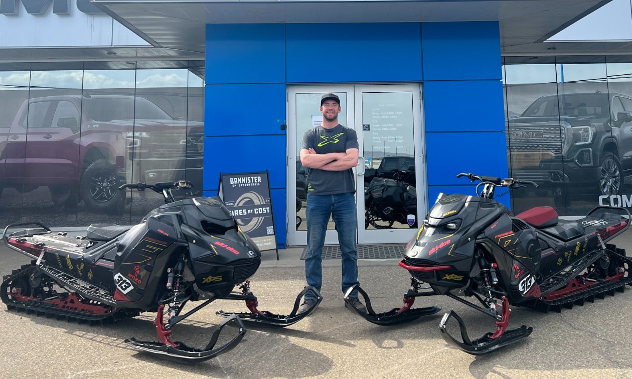 Brennan Mulvahill stands with his arms folded next to two 2023 Ski-Doo Freeride snowmobiles in front of a GM dealership. 