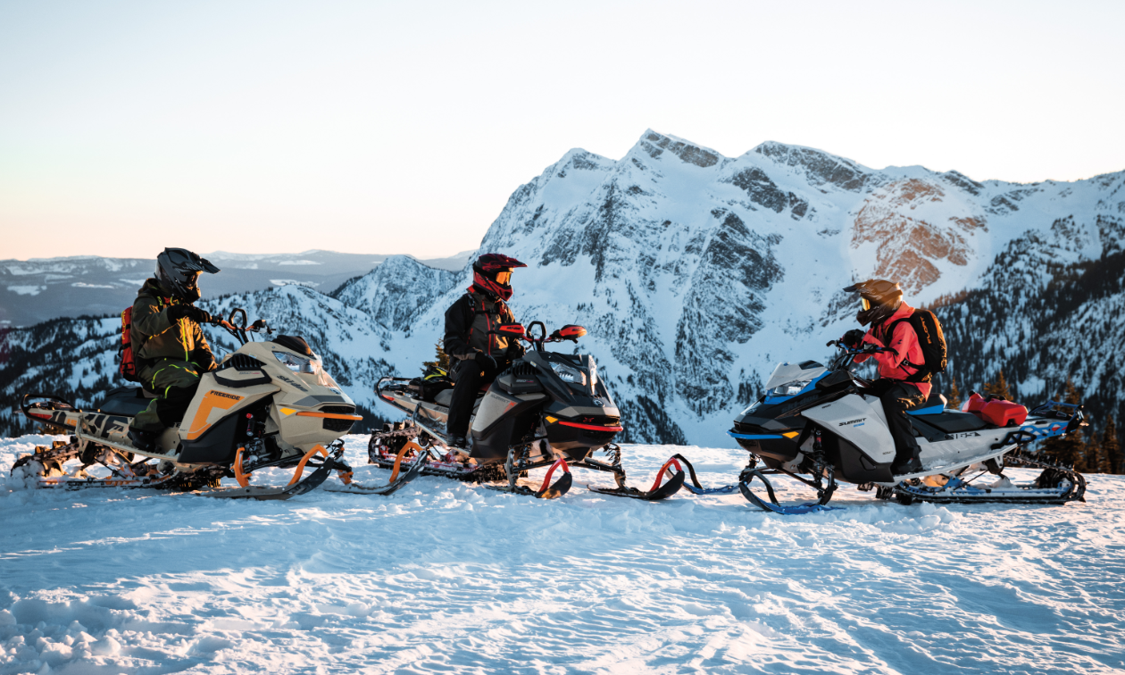 Three snowmobilers form a semi-circle on top of a snowy mountain. 