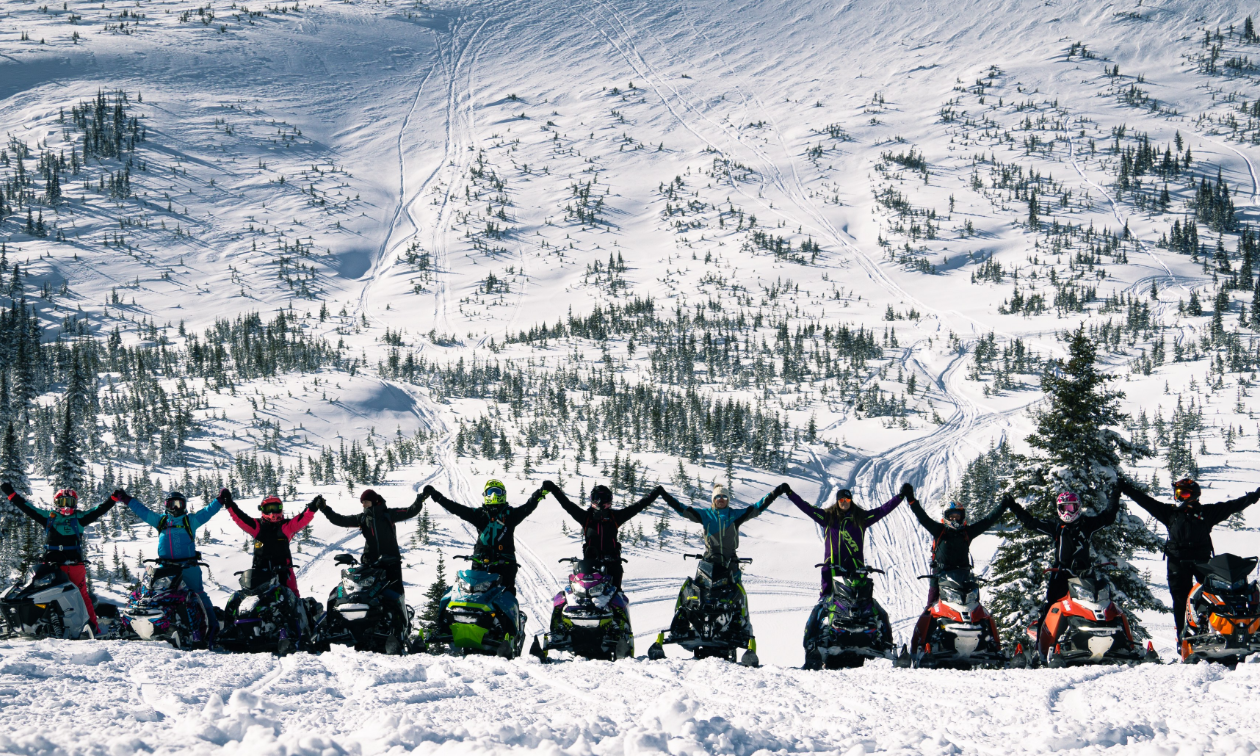 A row of female snowmobilers pose in a line with their arms outstretched, touching the rider next to them. 