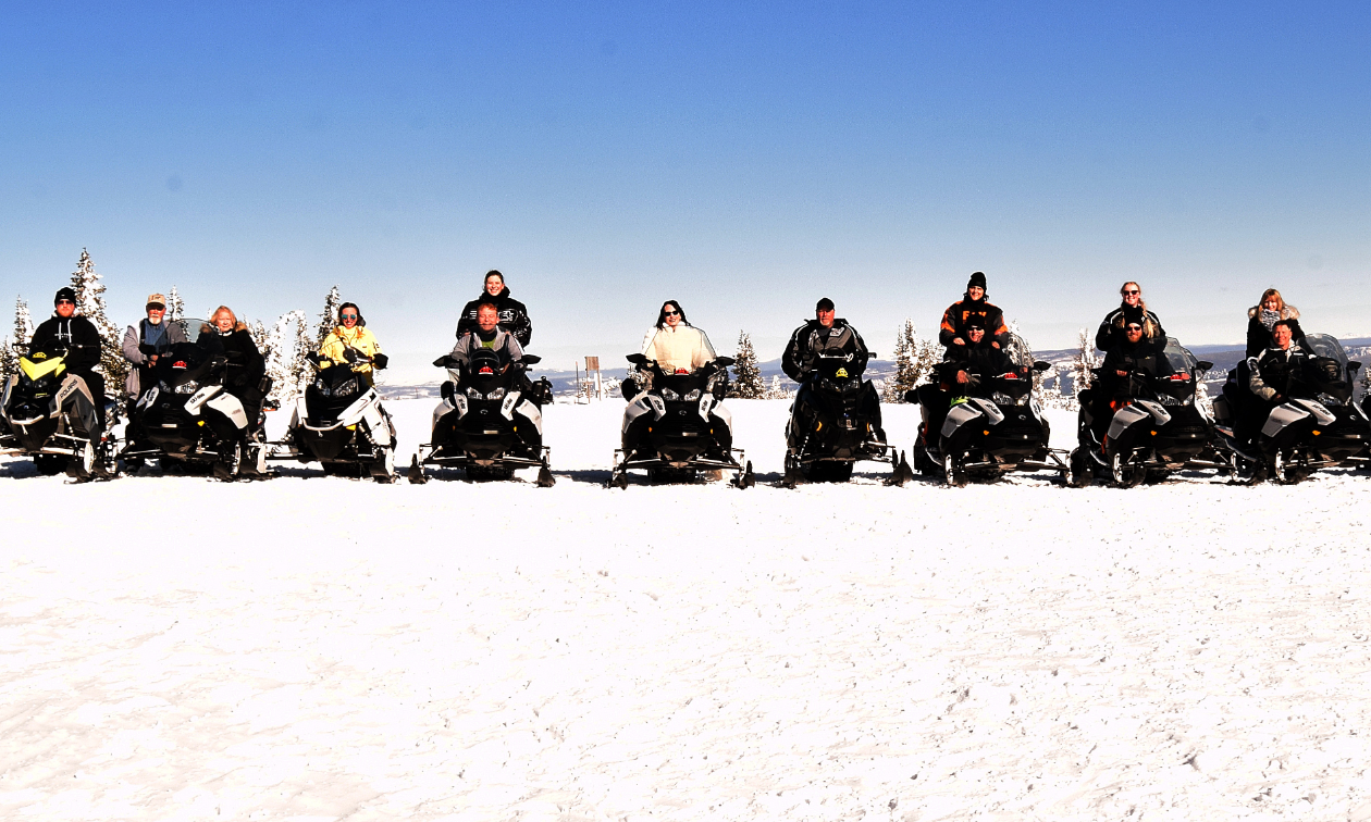 A lineup of snowmobilers on a mountain on a blue sky day. 