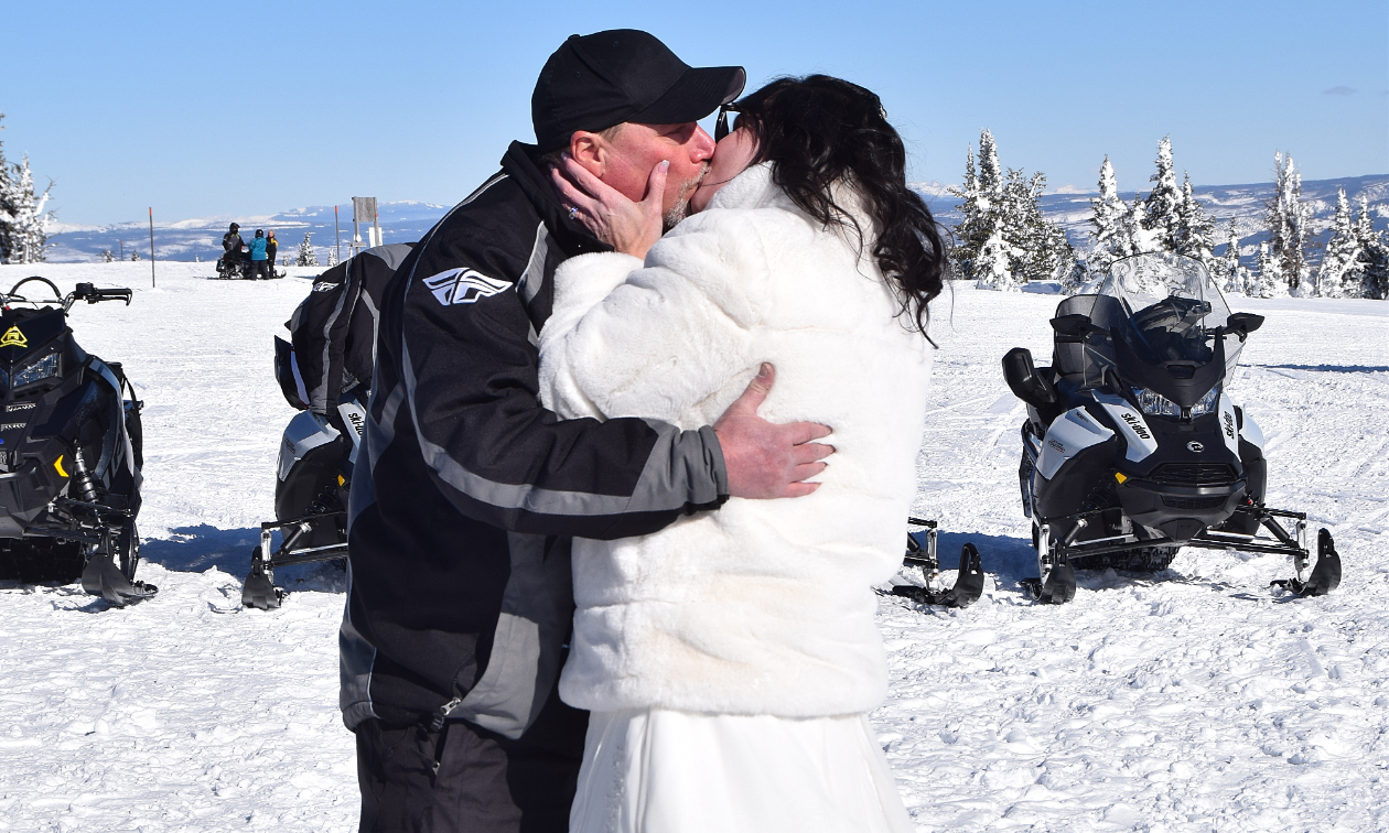 Denny and Chris Steigerwald kiss on top of a mountain with snowmobiles in the background.
