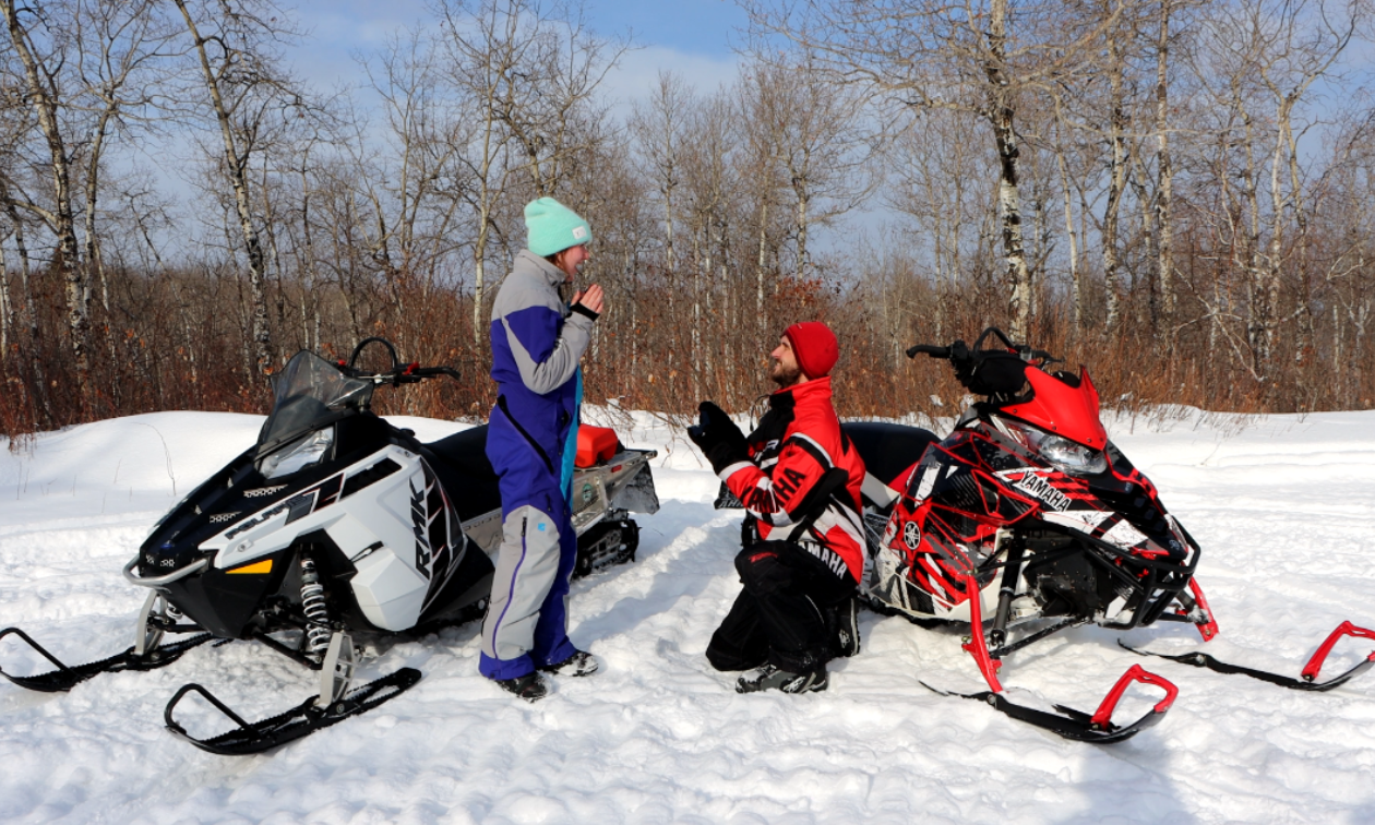 A man wearing red and black proposes to a woman wearing blue on a snowy trail next to snowmobiles. 