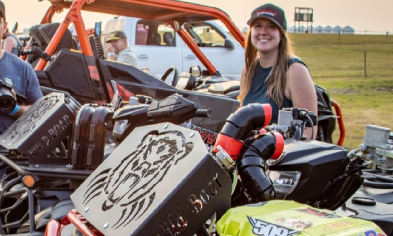Erin Kosowan smiles while wearing a baseball cap. She has long red hair and stands in the midst of ATVs. 