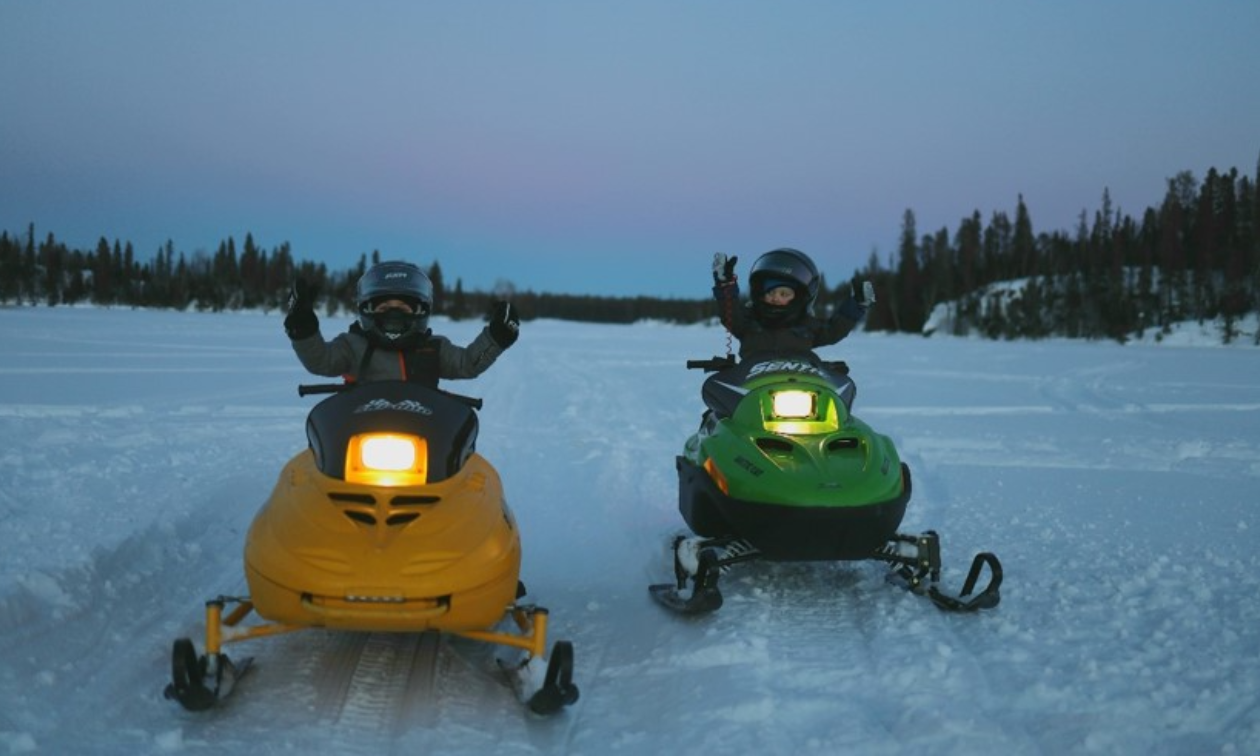 Two children ride small yellow and green snowmobiles next to each other on snow. 