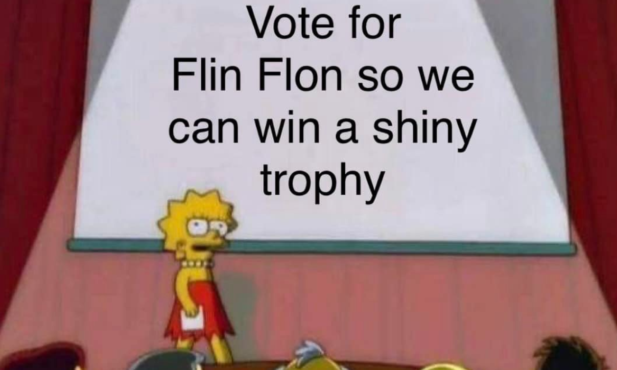 Lisa Simpson stands on a stage with a sign that says Vote for Flin Flon so we can win a shiny trophy.