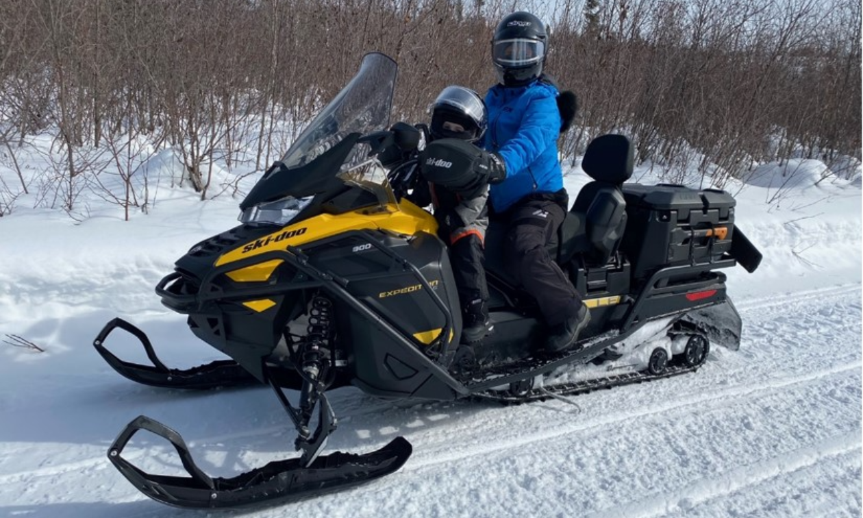 A black and yellow snowmobile with a man and child riding on it on a snowmobile trail.