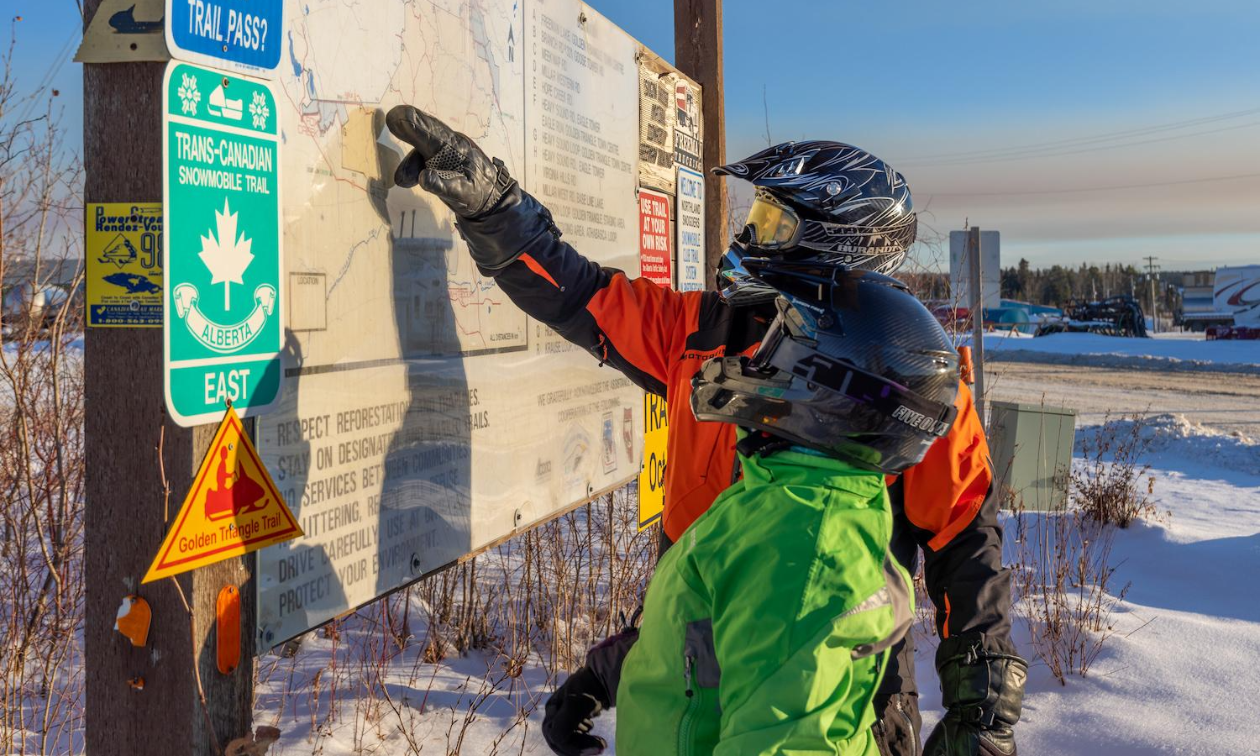 Two snowmobilers stand in front of a sign next to a snowmobile trail. One rider is pointing at something on the sign. 