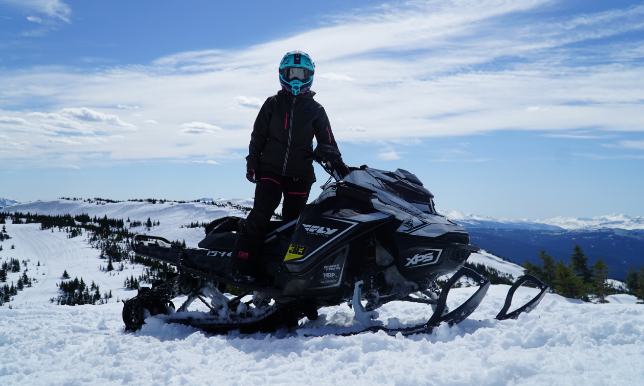 Genevieve Byl stands on her snowmobile at the top of a mountain.