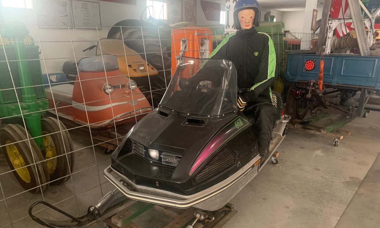 A mannequin sits on top of an old black vintage snowmobile in a warehouse.