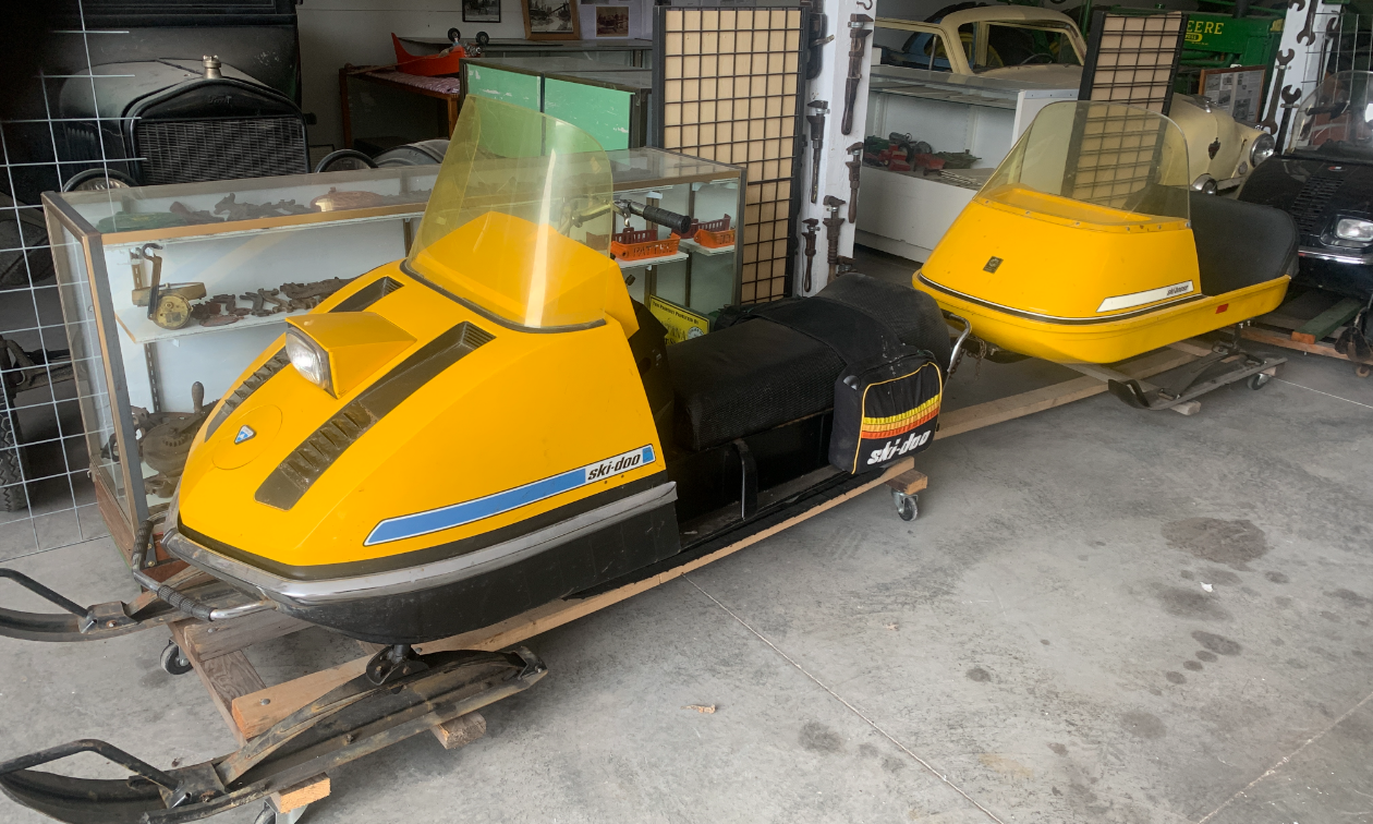 An old vintage yellow snowmobile with a small yellow trailer behind it. 