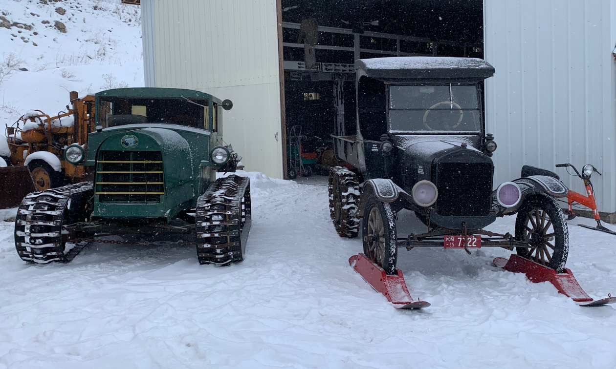 Old snow machines with tracks and skis are parked in front of a warehouse. 