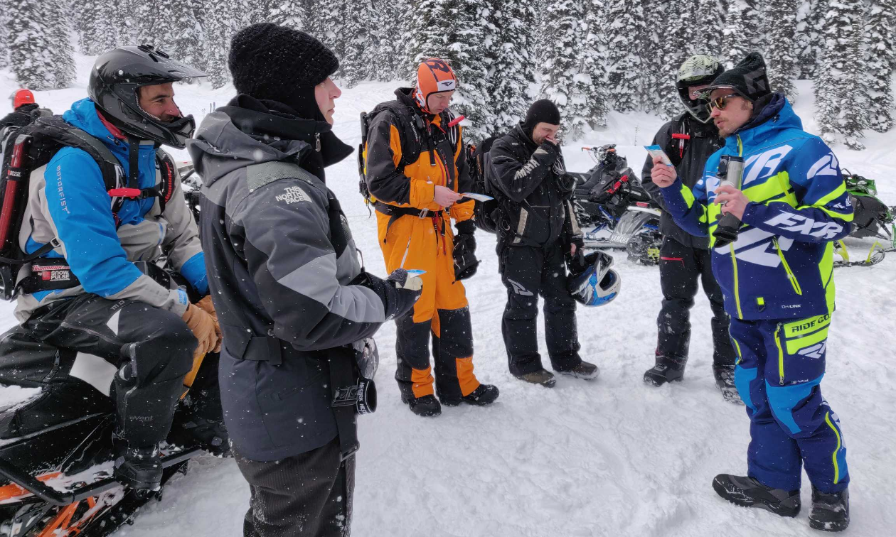 A group of snowmobilers stand in a circle while receiving instruction. 