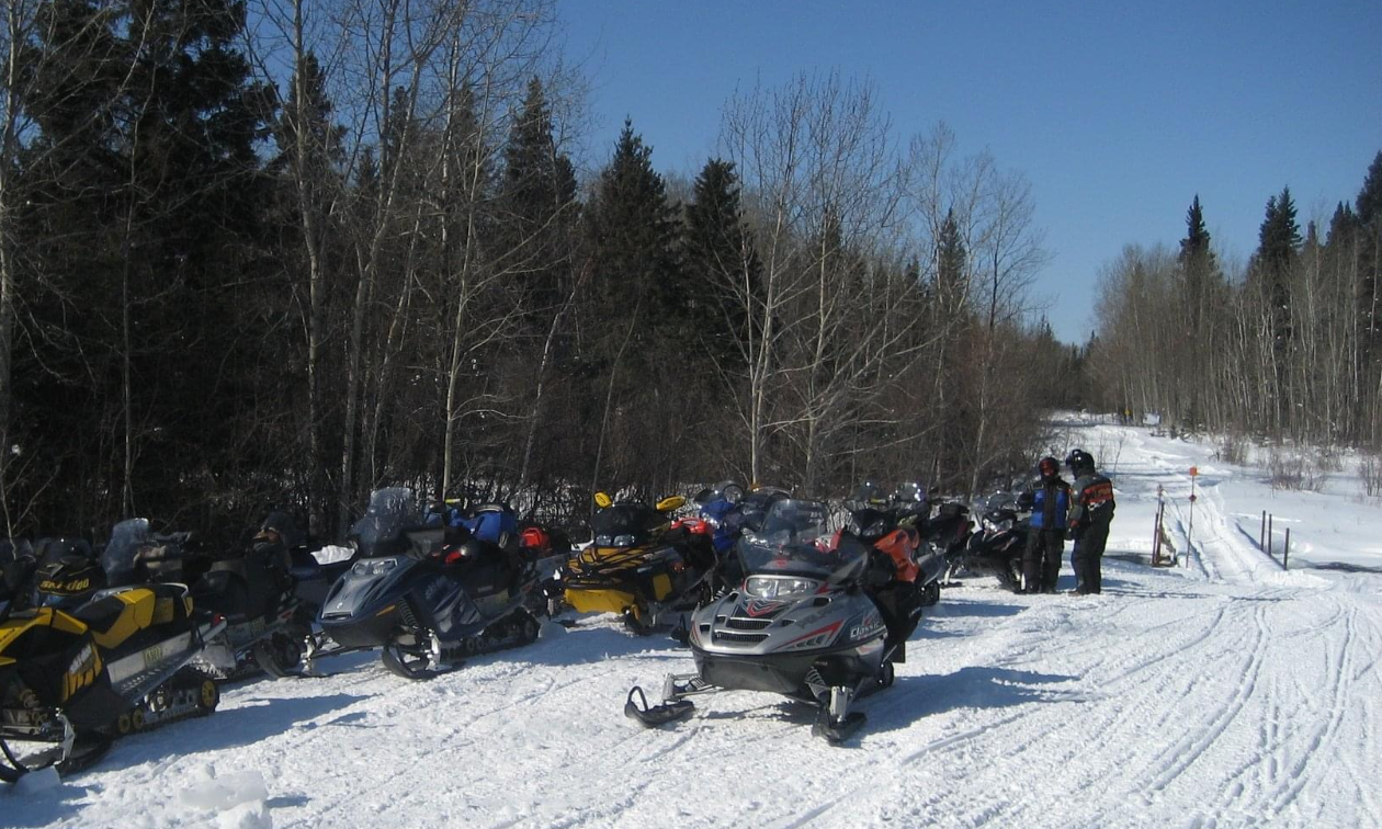 A line of snowmobiles parked on a long, groomed snowmobile trail. 