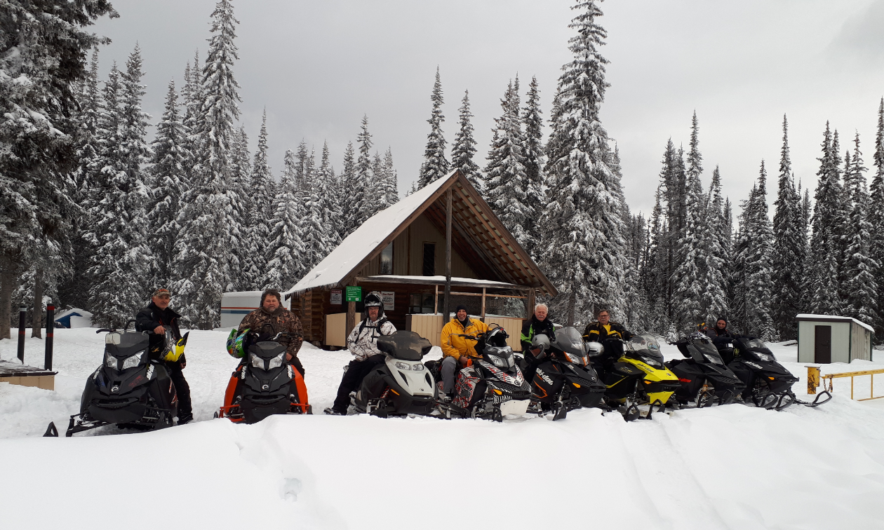 A row of snowmobilers line up in front of a tall cabin with a steep roof amidst the forest. 