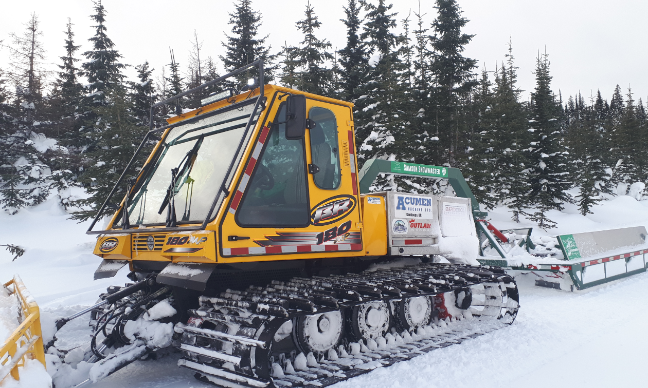 A large yellow snowmobile groomer pulls a drag behind it while clearing a trail. 