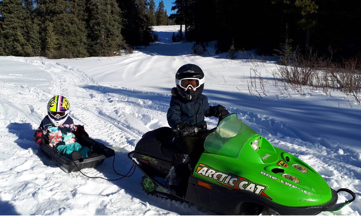 A boy riding a green snowmobile pulls a girl on a toboggan behind him. 