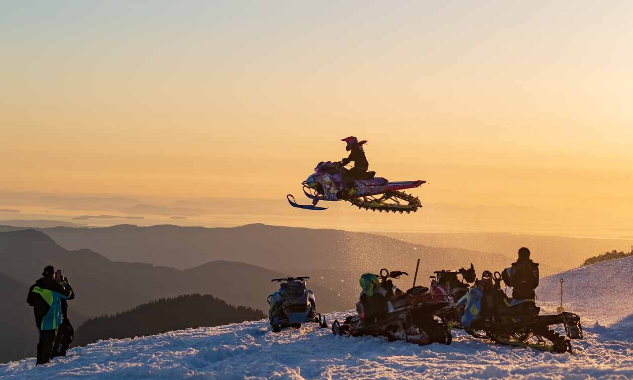 A snowmobile flies through the air, above a row of sledders underneath and looks like it’s higher than the mountains in the distance. An orange sunset provides dim but soothing lighting. 