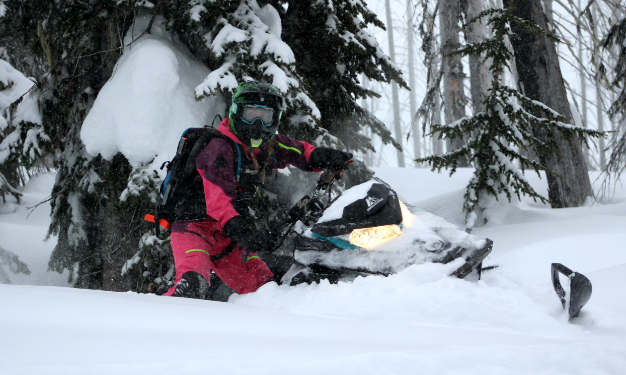 Kirsten Patton wears pink outerwear while driving her Ski-Doo snowmobile through powder between trees. 