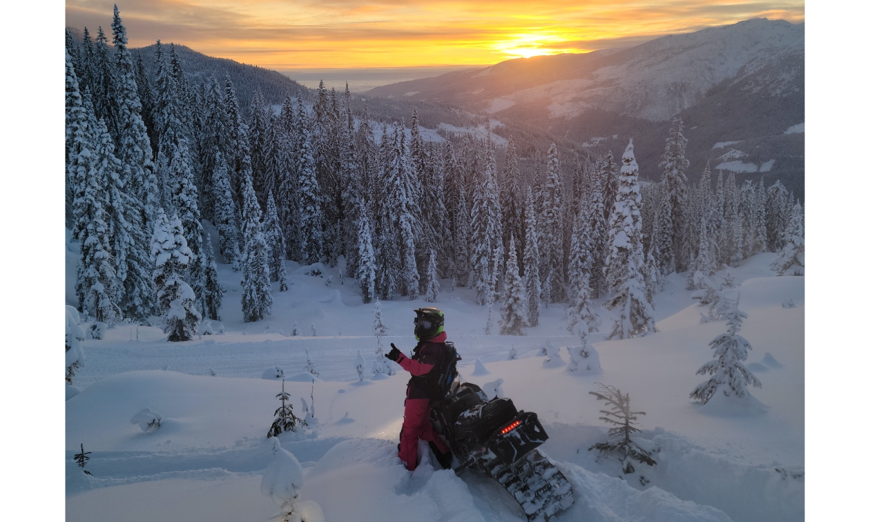 Kirsten Patton stands next to her Ski-Doo snowmobile while looking out to the sunset on the horizon of the distant mountains. 