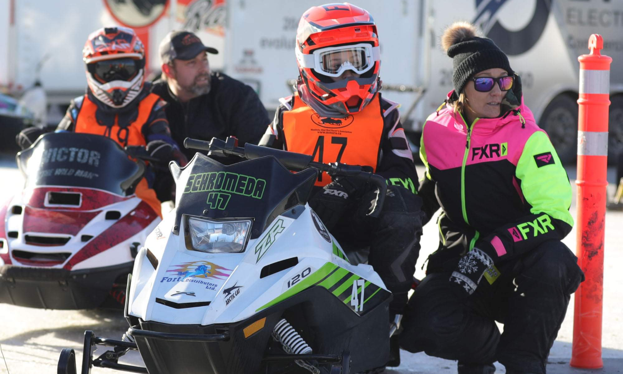Adults squat down next to kids riding snowmobiles in preparation for a race. 