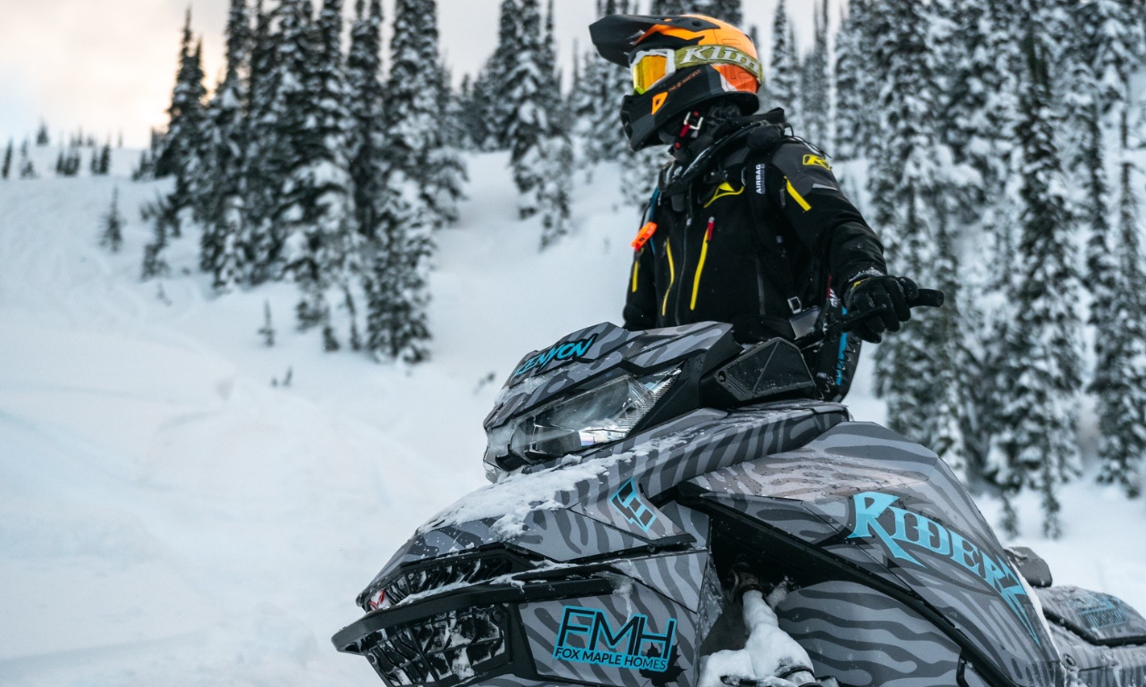 Mason Kenyon sits on a blue and black snowmobile wearing an orange helmet and orange goggles. 