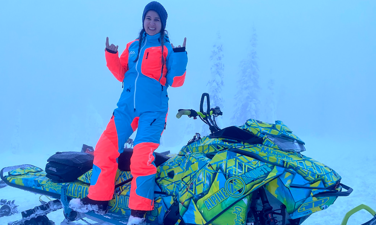 Pam DeRosa stands on her snowmobile, which has a neon green and blue sled wrap with the name Alpine Addicts written on the side. 